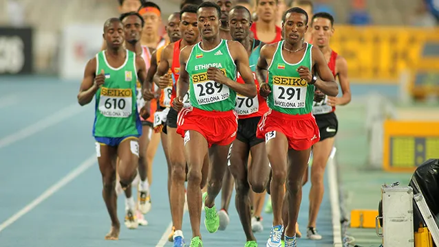 Fotografia di una gara di mezzofondo o fondo disputata durante i campionati di atletica leggera di Barcellona del 2012, con il marchio Seiko visibile sulla segnaletica a bordo pista. Si vede un folto gruppo di corridori maschi in piena corsa su una pista di atletica blu. In primo piano spiccano due atleti etiopi che indossano la divisa Adidas verde e rossa, con i numeri di gara 289 e 291. Dietro di loro sono visibili diversi altri concorrenti con divise di varie nazionali, tra cui il corridore numero 610.